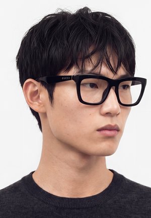 Giovane uomo con capelli corti neri che indossa grandi occhiali rettangolari neri Gucci, guarda leggermente verso destra, indossa un maglione scuro.
