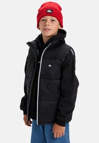 Pojke som bär en röd mössa, svart pufferväst över en svart hoodie och blå jeans, stående mot en enkel vit bakgrund.