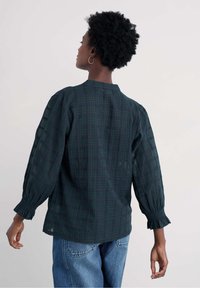 Žena s krátkymi kučeravými vlasmi, oblečená v tmavozelenej kockovanej blúzke s našitými rukávmi a vo svetle modrých džínsach, zobrazená zozadu na svetlom pozadí.