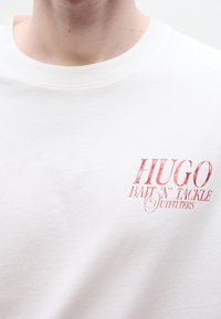 Άσπρο βαμβακερό t-shirt με τυπωμένο λογότυπο σε ροζ χρώμα που γράφει "HUGO BAIT 'N' TACKLE OUTFITTERS" στην αριστερή πλευρά του στήθους. Κοντά μανίκια.