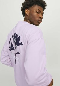 Sudadera de color púrpura claro hecha de material suave, con un diseño gráfico de acentos oscuros en la parte posterior, incluyendo texto y elementos florales.