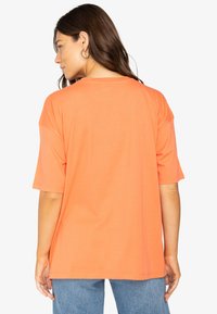 Short-sleeve, oversized t-shirt in bright orange fabric, featuring a round neckline and a smooth texture, paired with blue jeans.
