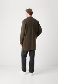 Olivgrön ullkappa med skräddarsydd design, utrustad med slagkrage och långa ärmar, kombinerad med mörka jeans och vita sneakers.
