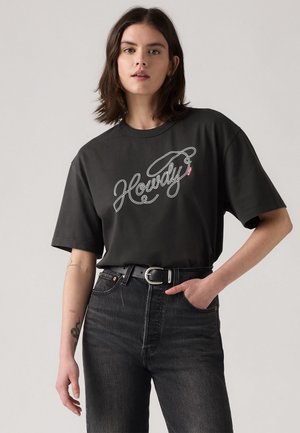 Sort t-shirt med korte ærmer, der har en hvid "Howdy" kursiv grafik. Sammen med højtaljede sorte denimjeans og et sort bælte.