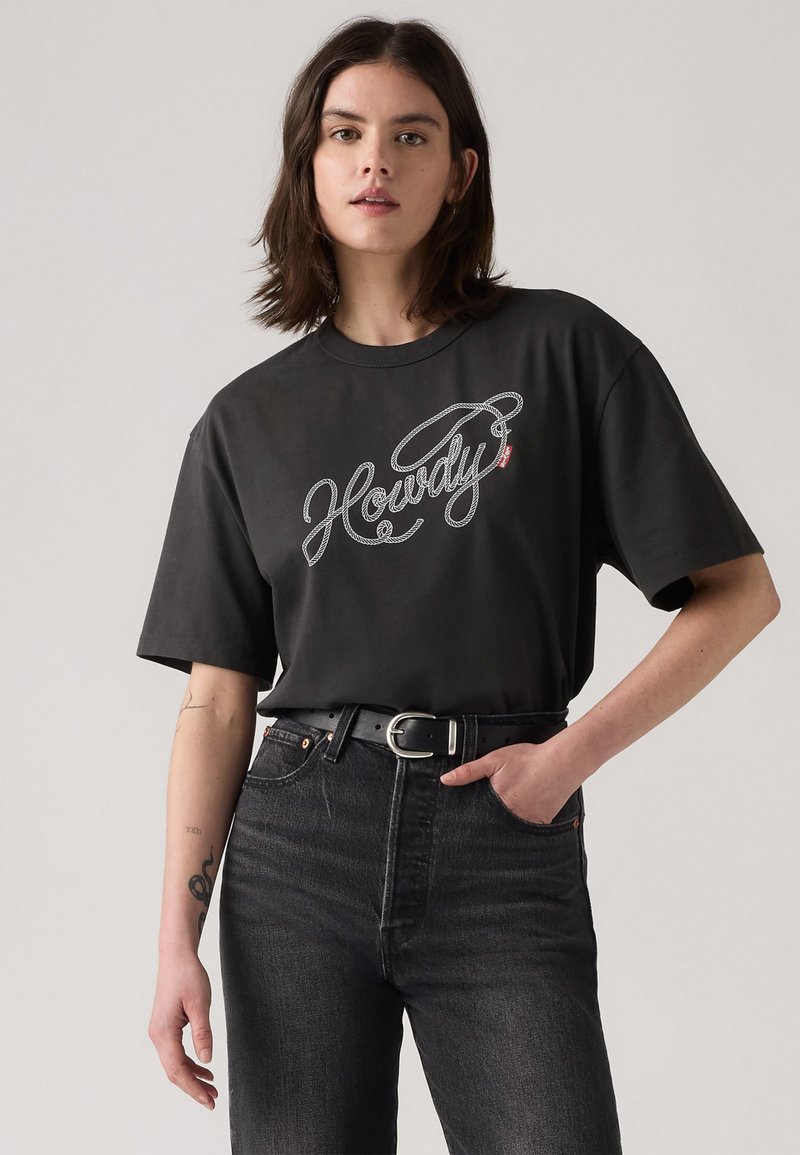 Camiseta negra de manga corta con un gráfico en cursiva blanco que dice "Howdy". Combinada con unos jeans de mezclilla negros de cintura alta y un cinturón negro.