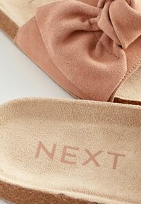 Světlé růžové semišové skluzné sandály s texturovanou mašlí a béžovou, texturovanou stélkou. Název značky "NEXT" v měkké růžové barvě.