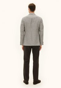 Mężczyzna stojący tyłem, ubrany w kraciasty blezer, czarne spodnie i czarne buty na prostym jasnym tle.