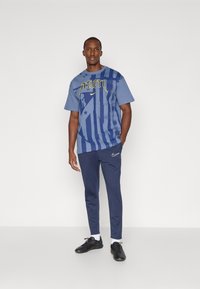 Blå grafisk t-shirt med abstrakte mønstre og tekst, parret med marineblå træningsbukser og sorte sneakers. Har Nike-logo på bukserne.