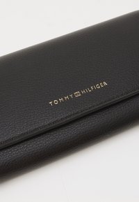 Black textured leather wallet with a gold "TOMMY HILFIGER" logo, featuring a slim rectangular design and subtle stitching detail.