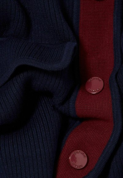 Tessuto a maglia di colore blu scuro con una striscia contrastante bordeaux, dotato di un bottone rotondo bordeaux. Il tessuto presenta una texture a coste e pieghe.
