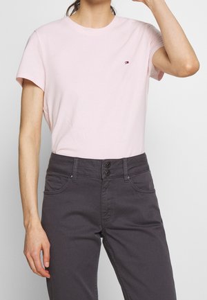Kvinde iført lyserød kortærmet t-shirt med lille logo på brystet og højtaljede mørkegrå bukser med to knapper.