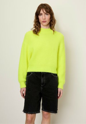 Felgroen gebreid sweater met een hoge hals en ribbelstructuur, gecombineerd met zwarte denimshorts met rafelige zomen en zakken.