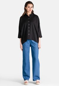 Camicia nera con bottoni, dal taglio comodo e colletto, abbinata a pantaloni a gamba larga blu chiaro, che mostrano un look casual ma contemporaneo.