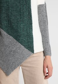 Pull en maille avec panneaux verts, blancs et gris ; section verte texturée, blanche lisse et manches grises côtelées. Design de ourlet asymétrique.