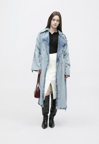 Mulher vestindo um casaco longo de ganga azul claro, top preto, saia branca com botões e fenda, botas pretas até ao joelho, e segurando uma mala de mão vermelha escura.