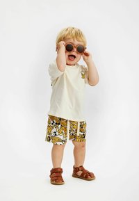 T-shirt en coton crème avec un graphisme de banane, shorts à motifs jaunes et noirs, sandales brunes à velcro, lunettes de soleil rondes marron.