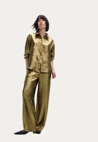 Conjunto de camisa metálica dorada y pantalones de pierna ancha con un acabado texturizado. La camisa cuenta con un cierre de botones y mangas enrolladas; los pantalones son de longitud hasta el suelo.