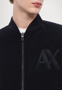 Giacca bomber nera con chiusura a zip, colletto a costine e logo "AX" ricamato sul petto. Tessuto liscio, vestibilità sartoriale.