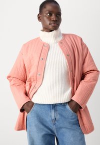 Rosa fodrad jacka med rund hals och tryckknappar, dragen över en vit ribbad polotröja, ihop med blå denimjeans.