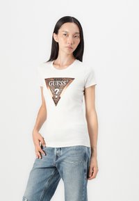 Hvid kortærmet t-shirt med et trekantet brunt grafisk motiv med teksten "GUESS®" og et spørgsmålstegn, parret med blå denimbukser.