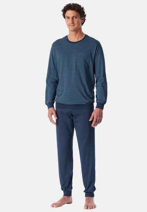 Mann steht barfuß und trägt einen passenden blauen Langarmtop und Hose mit gerippten Bündchen vor einem einfarbigen Hintergrund.