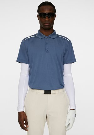 Lichtblauw poloshirt met witte accenten, korte mouwen en knopen; gecombineerd met beige broeken en een witte golfhandschoen. Zonnebril is gedragen.