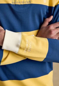Personne croisant les bras, portant un sweat-shirt à rayures bleues et jaunes avec "Joules MCMLXIX" brodé sur le poignet de la manche.