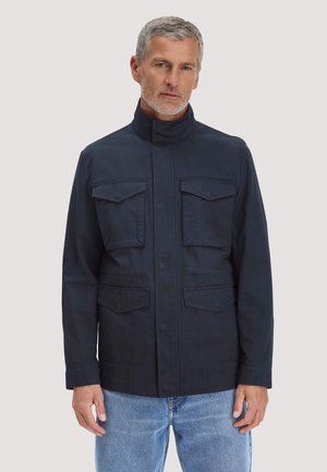 Middelbare man met kort grijs haar draagt een marineblauwe jas met vier klepzakken aan de voorkant en lichtblauwe jeans tegen een effen achtergrond.