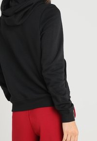 Svart hoodie med mjuk textur, utrustad med stor huva och ribbade muddar. Bärs med röda åtsittande byxor som visar en avslappnad passform.