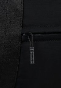 Schwarze Reißverschlusstasche aus strukturiertem Stoff und glattem Material, mit einem schwarzen Ziehband, das ein Diamantmuster und ein Logodetail zeigt.