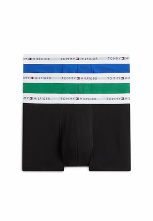 Joven sonriendo, de pie descalzo, vistiendo calzoncillos tipo bóxer azul brillante con cintura blanca etiquetada Tommy Hilfiger.