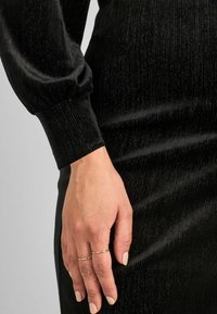 Fekete ruha texturált, bordás mintával. Hosszú ujjú, ráncolt mandzsettával. Két aranygyűrű a kezén, világos színű körmökkel.