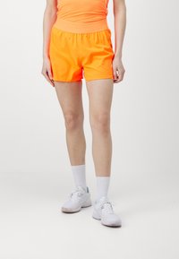 Neon oranssi urheilushortsit, joissa on joustava vyötärö, sileä pinta ja tiukka muotoilu. Yhdistetty valkoisiin tennareihin ja sukkiin.