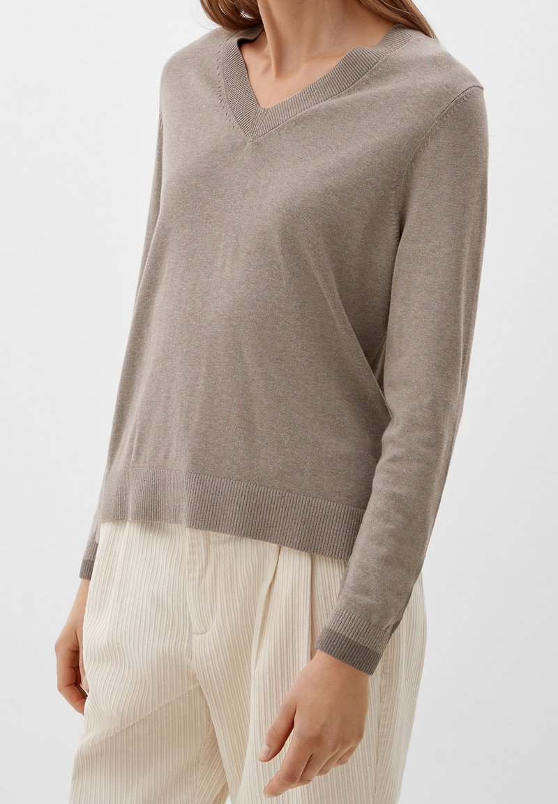 Pull beige en maille avec un col en V, coupe slim, et poignets et ourlet côtelés. Associé à un pantalon large texturé de couleur claire.