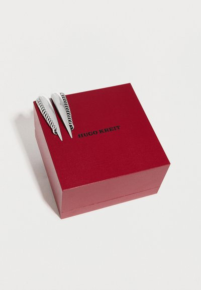 Κόκκινο τετράγωνο κουτί με λείο φινίρισμα, στο οποίο αναγράφεται το κείμενο "HUGO KREIT" με μαύρα γράμματα, και στην κορυφή έχει δύο ασημένια μεταλλικά κλιπ με σπειροειδείς λεπτομέρειες.