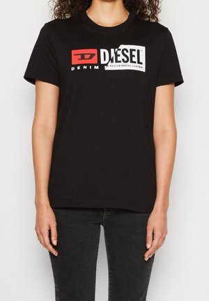 Person, die ein schwarzes Diesel T-Shirt mit rotem und weißem Logo-Text "DIESEL Denim for successful living" und dunklen Jeans trägt, steht vor einem schlichten Hintergrund.