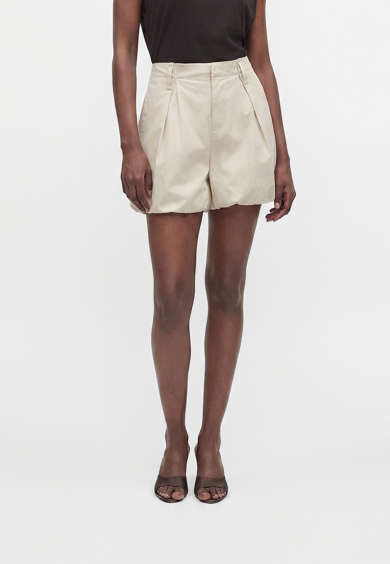 Personne portant un short plissé beige taille haute, un haut noir sans manches et des talons noirs ouverts à l'avant, debout devant un fond clair uni.