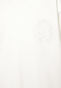 Maglietta bianca in cotone con un logo circolare testurizzato e in rilievo, accompagnato dalle scritte "COMUNITÀ" e "CULTURA", con accenti di foglie di alloro.