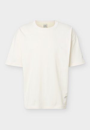 Tricou simplu alb, cu mâneci scurte și guler rotund, cu croială lejeră și un mic logo discret aproape de tivul de jos, pe un fundal deschis.