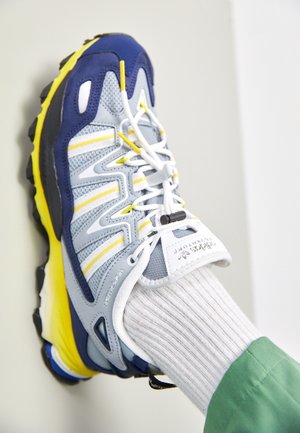 Chaussure de sport bleue, jaune et grise avec des lacets blancs portée avec une chaussette blanche côtelée et un pantalon vert sur un fond clair.