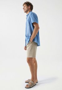 Camisa de botones azul claro con un bolsillo, pantalones cortos beige y sandalias marrones con dos correas de hebilla. El modelo está de lado contra un fondo plano.