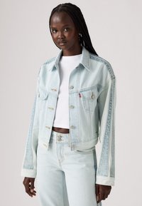 Chaqueta de mezclilla azul claro con un diseño cortado, que presenta botones dorados, dos bolsillos en el pecho y un ajuste ligeramente holgado. Tejido texturizado.
