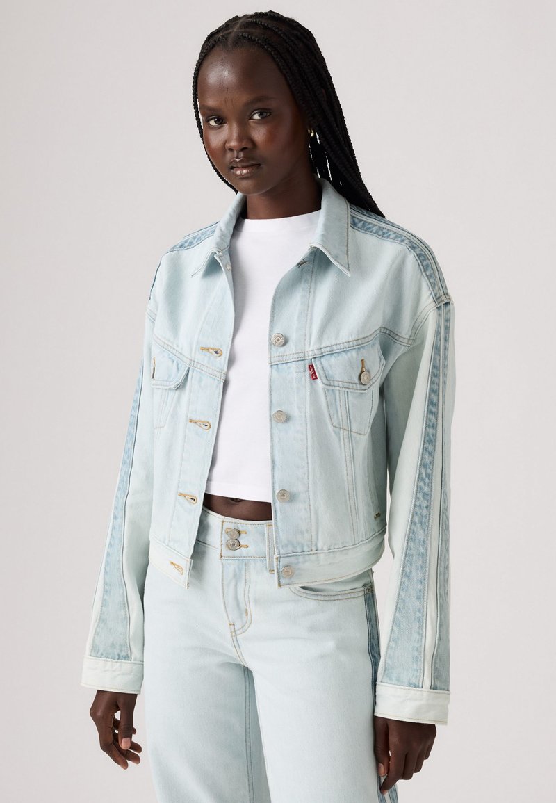 Chaqueta de mezclilla azul claro con un diseño cortado, que presenta botones dorados, dos bolsillos en el pecho y un ajuste ligeramente holgado. Tejido texturizado.