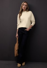 Sweat-shirt couleur crème avec poignets côtelés, jean évasé bleu foncé, bottines en daim beige et sac fourre-tout camel. Fond uni.