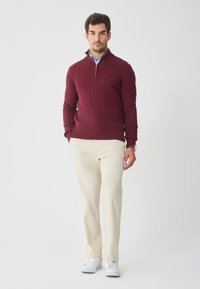 Maglione a coste color bordeaux con collo a mezza zip, abbinato a pantaloni chiari e sneakers bianche, caratterizzato da un tessuto testurizzato.