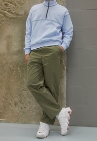 Světle modrý fleecový pulovr s polovičním zipem a přední kapsou, spárovaný s olivově zelenými kalhotami a bílými teniskami s červenými akcenty.