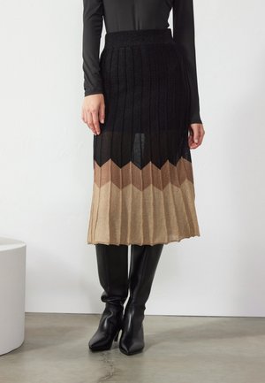 Kobieta ubrana w czarną bluzkę z długim rękawem, plisowaną spódnicę z czarno-brązowo-beżowym wzorem w zygzak oraz czarne kozaki za kolano, stojąca w pomieszczeniu.