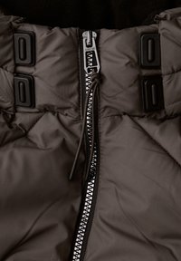 Veste matelassée marron avec fermeture éclair avant, bouton à bascule et attaches en plastique noir. Texture douce et design isolant visibles.