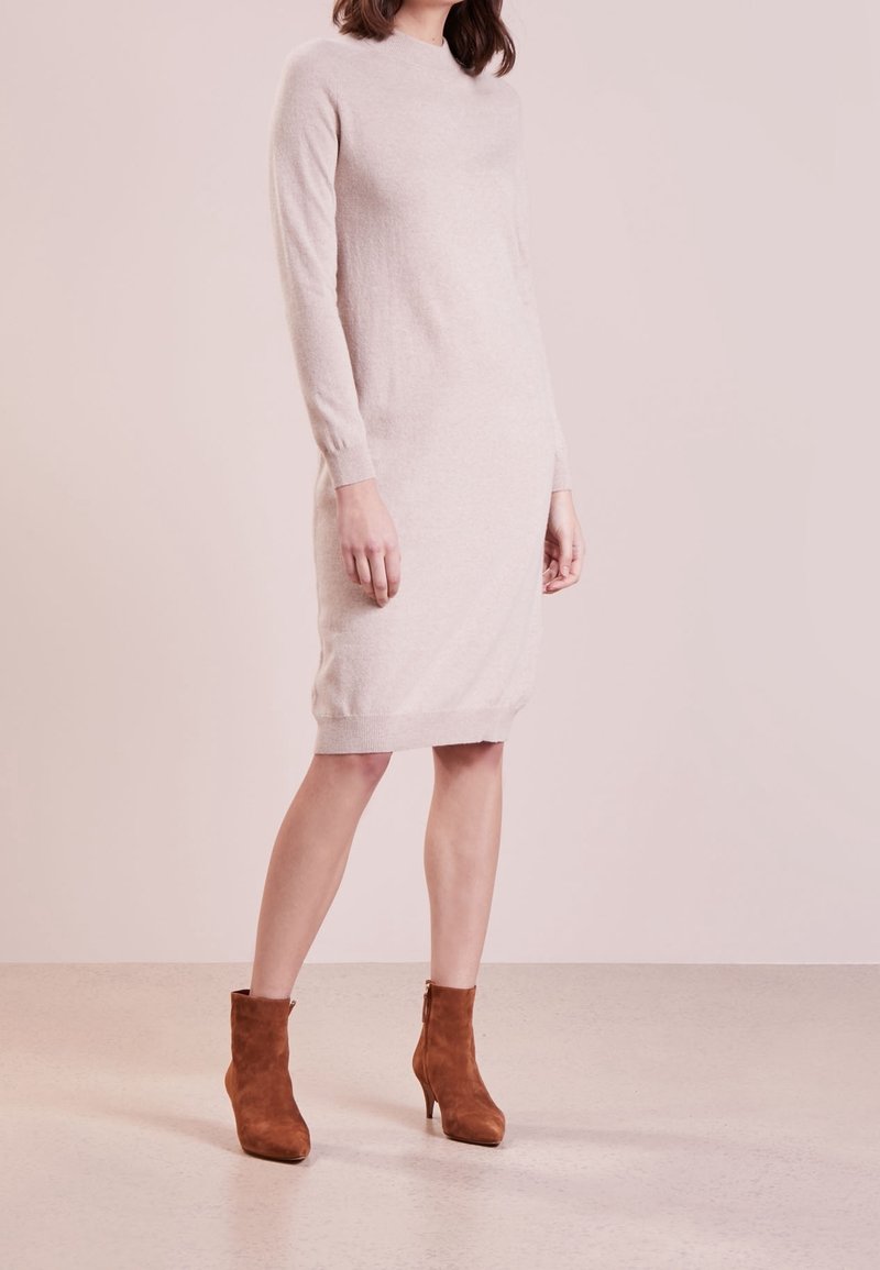 Femme portant une robe-pull beige clair à manches longues, longueur genou, associée à des bottines en daim marron, debout sur un fond neutre.