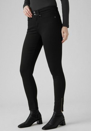 Femme portant un pantalon noir ajusté avec des fermetures éclair aux chevilles et des bottines noires, debout devant un fond uni.